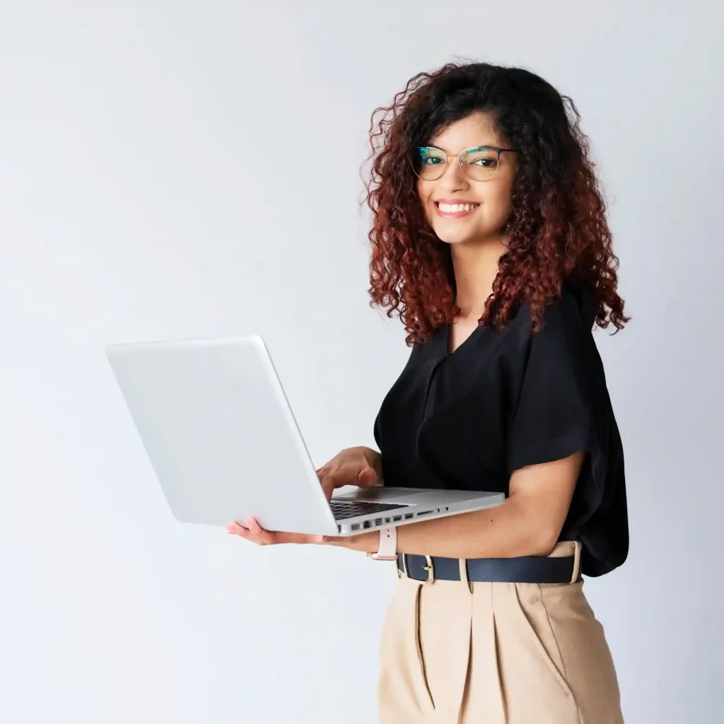 A young woman with curly hair wearing glasses and a black blouse is smiling while holding an open laptop in grace academy