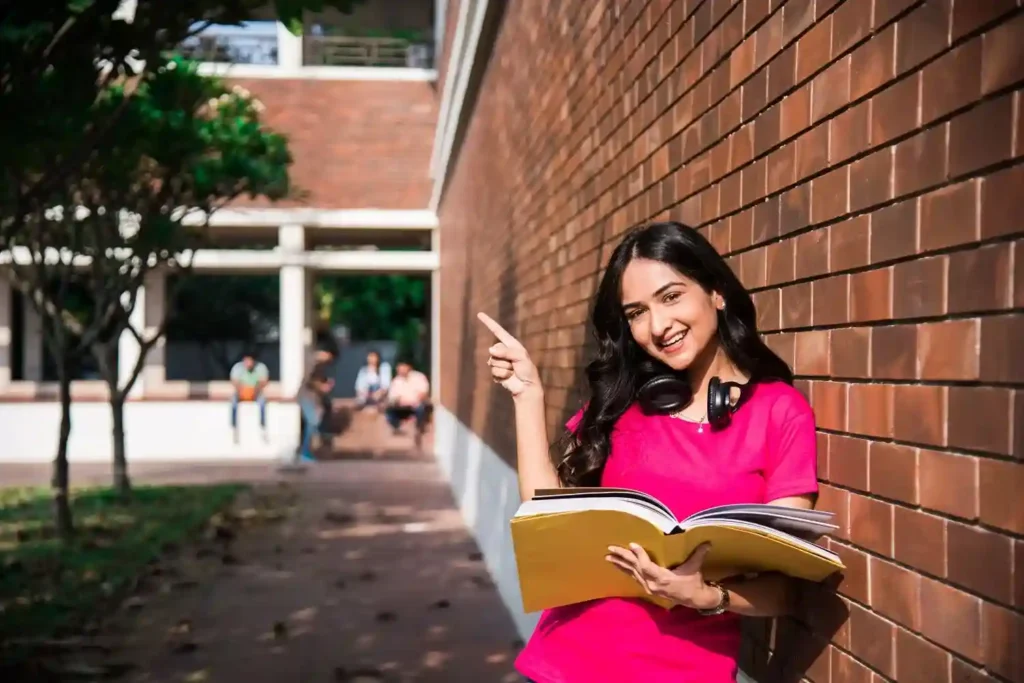 Smiling young alumina of grace academy in a pink top holding an open book and pointing while leaning against a brick wall on a college campus.