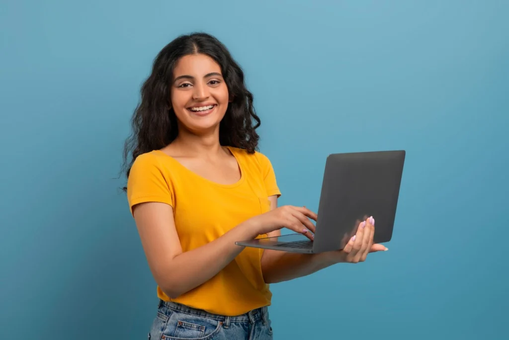 A young woman smiling while holding a laptop and typing, wearing a yellow t-shirt against a blue background.
