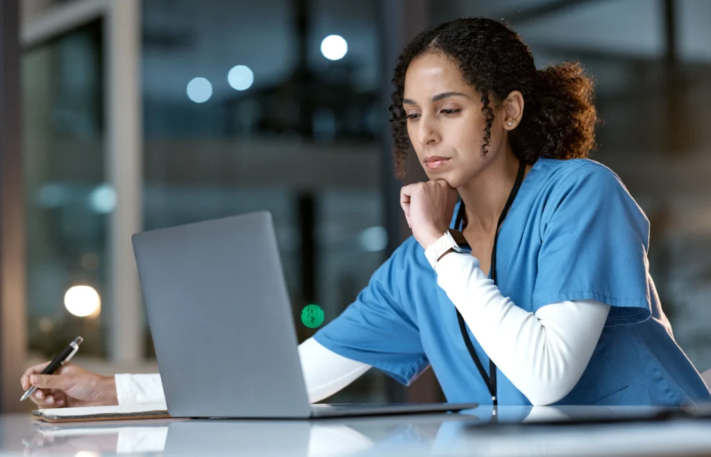 Nurse in blue scrubs working on a laptop at night in a hospital while taking notes in a notebook. OET Exam for Nurses